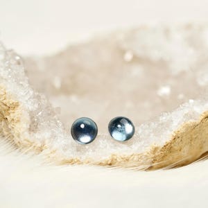 May include: Two small, round, blue-grey stud earrings. The earrings are displayed on a white, textured surface, possibly a geode, against a soft, white background. The earrings have a glossy finish.