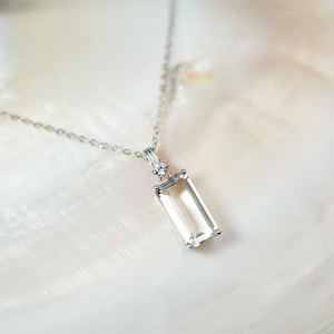 May include: A silver necklace with a rectangular, clear gemstone pendant. The pendant is suspended from a delicate chain and is set with small, clear accent stones. The necklace is displayed on a white, textured surface.