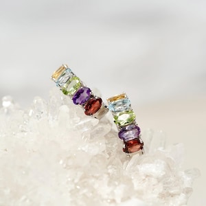 May include: A pair of silver earrings, each featuring a row of colorful gemstones. The gemstones include yellow, blue, green, purple, and red hues. The earrings are displayed on a white, crystalline surface.