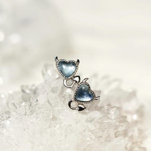 May include: A pair of silver-toned earrings featuring heart-shaped charms with small devil horns and a pointed tail. Each heart contains a translucent, light blue stone set within a beaded metal border, connected by a small silver link.