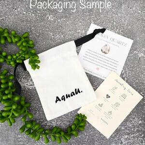 May include: A white drawstring bag with the word "Aquah." printed in black. The bag is sitting on a grey surface with a green plant. A card with the text "Smokey Quartz for grounding & relaxation" is next to the bag.
