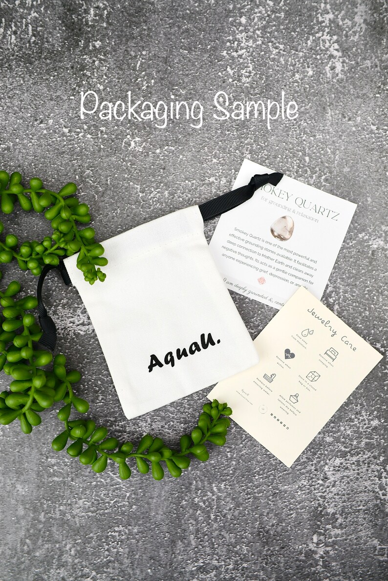 May include: A white drawstring bag with the word "Aquah." printed in black. The bag is sitting on a grey surface with a green plant. A card with the text "Smokey Quartz for grounding & relaxation" is next to the bag.