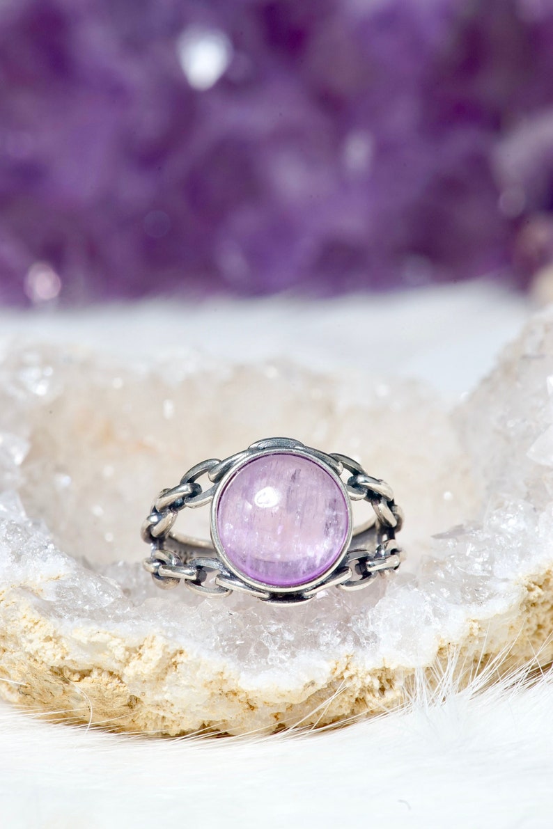 May include: A silver ring featuring a light purple gemstone set within a chain-link design. The ring is displayed on a white and beige surface.