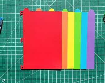 Può includere: Una collezione di divisori per cartelle colorati in rosso, arancione, giallo, verde, blu e viola. Tre penne arancioni, viola e blu a sinistra e una penna verde a destra. Gli oggetti sono su un tappetino da taglio verde.