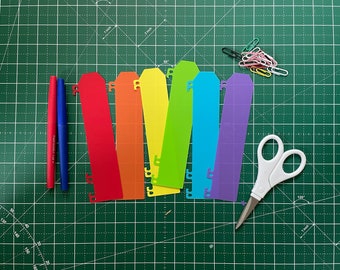 Può includere: Un set di sette divisori di carta arcobaleno con fori per un raccoglitore. I divisori sono disposti in un arcobaleno, con rosso, arancione, giallo, verde, blu, indaco e viola. I divisori sono su un tappetino da taglio verde con un paio di forbici bianche.