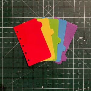 Może przedstawiać: Pięć kolorowych kartek indeksowych ze zaokrąglonymi rogami i dziurkowaniem po lewej stronie. Kartki są czerwone, żółte, zielone, niebieskie i fioletowe.