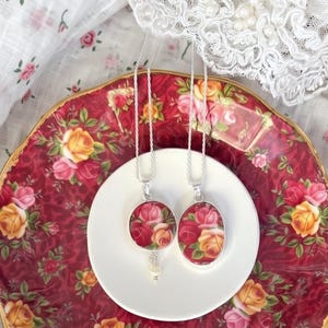 Può includere: Due collane con ciondoli in argento con disegni floreali poggiano su un piatto bianco, posto su un piatto rosso floreale. I ciondoli hanno una forma rotonda e ovale con un motivo a rose nei toni del rosso, rosa e giallo. Il piatto rosso ha un bordo dorato.