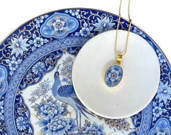 Może przedstawiać: Serwowana misa z niebiesko-białej porcelany z pozłacanym wykończeniem i wzorem pawia. Misa ma mniejszą, białą, okrągłą misę w środku. Złoty łańcuszek z niebiesko-białym porcelanowym wisiorkiem zwisa ze środkowej misy.