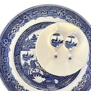 May include: A pair of silver earrings with a blue and white boat design, displayed on a small white plate atop a larger blue and white patterned plate. The earrings have dangling beads. The plates have a classic blue and white design.