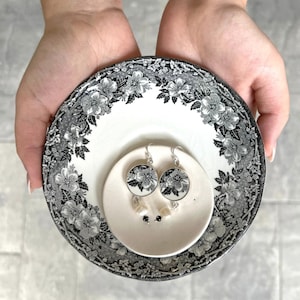 May include: A pair of black and white floral earrings with silver hooks and white pearl accents on a white saucer with a black and white floral pattern.