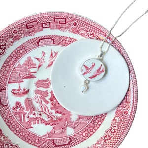 Puede incluir: Un collar de plata con un colgante de porcelana blanca con un diseño floral rojo y blanco. El colgante está colgado de un platillo blanco con un diseño floral rojo y blanco.