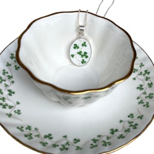 Puede incluir: Un collar de cadena de plata con un colgante ovalado blanco con un diseño de trébol verde. El colgante está sentado en una taza de té blanca con un borde dorado.