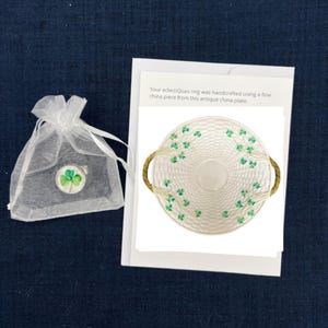 Op de afbeelding: Een wit porseleinen bord met een geweven ontwerp en groene klaveraccenten. Het bord heeft twee handvatten en is versierd met klavers. De tekst "Your eclectiQuas ring was handcrafted using a fine china piece from this antique china plate." is afgedrukt op een witte kaart.