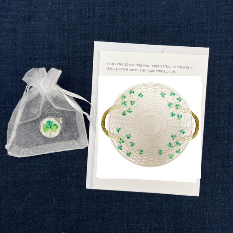 Op de afbeelding: Een wit porseleinen bord met een geweven ontwerp en groene klaveraccenten. Het bord heeft twee handvatten en is versierd met klavers. De tekst "Your eclectiQuas ring was handcrafted using a fine china piece from this antique china plate." is afgedrukt op een witte kaart.