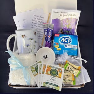 Puede incluir: Una caja de regalo llena de art&iacute;culos de cuidado personal. Incluye una taza blanca con dise&ntilde;os de abejas y flores, un cuenco blanco peque&ntilde;o con una "M", bolsitas de t&eacute;, pastillas, paquetes de Propel, un pa&ntilde;o y tarjetas inspiradoras. La caja est&aacute; atada con una cinta azul claro.