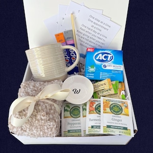 Peut inclure: Un coffret cadeau de soins personnels. Il comprend une tasse cr&egrave;me, une petite soucoupe blanche avec un "W", des sachets de th&eacute;, des pastilles et une couverture beige clair nou&eacute;e avec un ruban cr&egrave;me. Des cartes avec des messages inspirants sont &eacute;galement incluses.