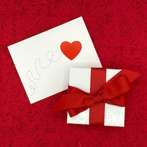 May include: A white card with a red heart and a dotted line that says "I love you". A white gift box with a red ribbon is in the foreground.
