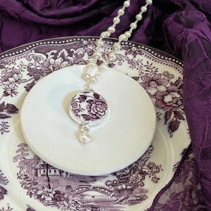Puede incluir: Un collar de plata con un colgante floral en tonos blancos y morados, con una cadena de perlas y una sola perla colgante. El colgante está sobre un plato de cerámica blanco con un estampado floral, sobre un fondo morado.