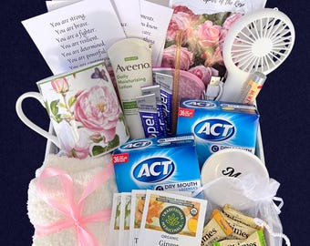 Pode incluir: Uma caixa de presente contendo uma caneca floral, loção Aveeno, duas caixas de pastilhas ACT para boca seca, um ventilador de mão branco, protetor labial Burt's Bees, um pequeno prato branco com a letra M, chá de gengibre e balas de gengibre.