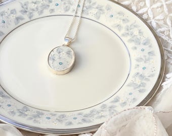 Może przedstawiać: Srebrny naszyjnik z wisiorkiem z owalną ceramiczną wkładką z delikatnym jasnoniebieskim i szarym wzorem kwiatowym. Wisiorek wisi na cienkim srebrnym łańcuszku, spoczywając na białym porcelanowym talerzu z pasującą kwiatową obwódką.