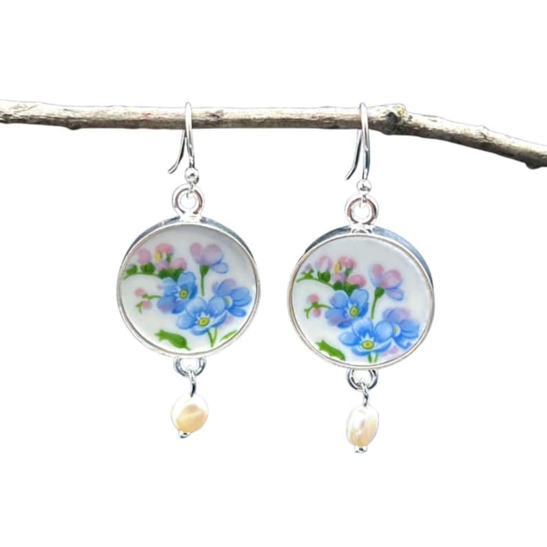 Pode incluir: Um par de brincos prateados. Cada brinco apresenta um disco redondo de porcelana branca com um desenho floral de flores azuis e rosas. Uma pequena p&eacute;rola creme pende da parte inferior de cada brinco. Os brincos est&atilde;o pendurados num ramo fino e castanho.