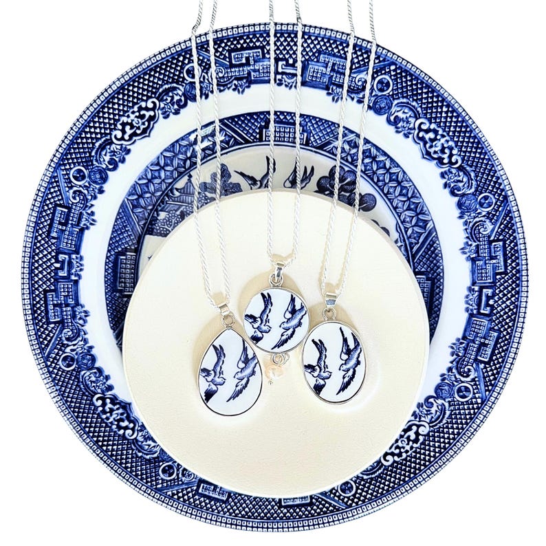 May include: Three silver necklaces with blue and white porcelain pendants featuring a bird design. The necklaces are displayed on a blue and white porcelain plate.