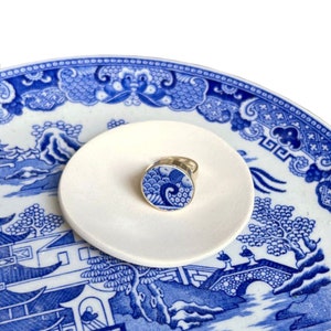 Può includere: Un anello d'argento con una faccia rotonda in porcellana blu e bianca con un motivo a onde. L'anello è esposto su un piattino rotondo bianco, sullo sfondo di un piatto a motivi blu e bianchi.