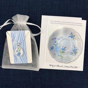 Pode incluir: Um colar de prata com um pingente floral, apresentado em um saco de presente branco transparente. O pingente apresenta flores azuis em um fundo branco. Um cart&atilde;o mostra um prato de porcelana correspondente com o texto "Royal Albert, Forget Me Not."