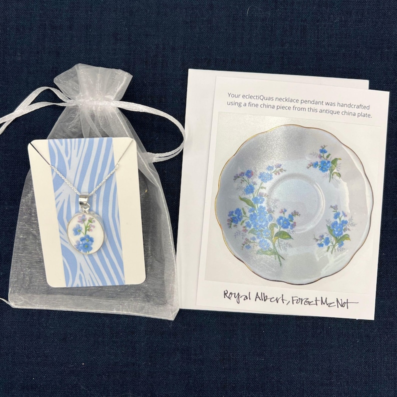 Pode incluir: Um colar de prata com um pingente floral, apresentado em um saco de presente branco transparente. O pingente apresenta flores azuis em um fundo branco. Um cart&atilde;o mostra um prato de porcelana correspondente com o texto "Royal Albert, Forget Me Not."
