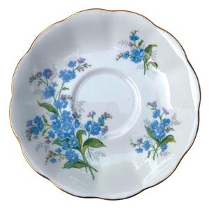 Pode incluir: Pires branco com borda ondulada e acabamento dourado. &Eacute; decorado com buqu&ecirc;s de flores azuis de mios&oacute;tis e folhas verdes. O centro tem uma &aacute;rea rebaixada para uma x&iacute;cara de ch&aacute;.