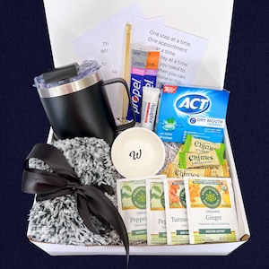 Peut inclure: Coffret cadeau de soins personnels. Il comprend un mug noir, un petit bol blanc avec un "w", des sachets de th&eacute;, des pastilles et une &eacute;charpe grise duveteuse nou&eacute;e avec un ruban noir. Contient aussi un tube d'Aquaphor et des sachets Propel.