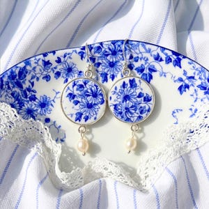 Op de afbeelding: Een paar ronde oorbellen met een blauw-wit bloemmotief op een keramiekachtig oppervlak, gezet in zilverkleurig metaal. Elke oorbel hangt aan een zilveren haakje en is versierd met een kleine, crèmekleurige parel.