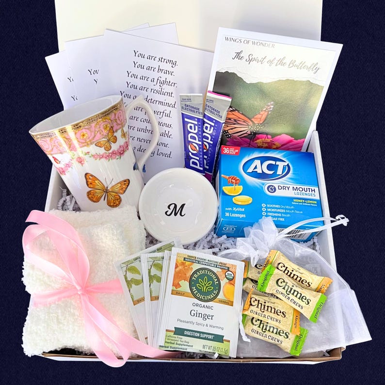Puede incluir: Una caja de regalo llena de varios art&iacute;culos, incluyendo una taza floral con mariposas, un cuenco blanco peque&ntilde;o con la letra "M", bolsitas de t&eacute;, caramelos de jengibre y un folleto titulado "Wings of Wonder". Una cinta rosa adorna un pa&ntilde;o blanco.