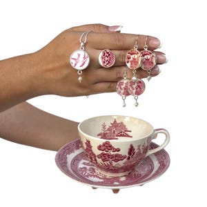 May include: A collection of red and white jewelry, including a necklace, ring, and earrings, displayed with a teacup and saucer. The jewelry features a circular design with a pearl accent. The teacup and saucer have a matching red and white pattern.
