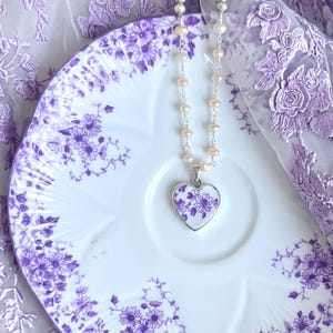 Op de afbeelding: Een ketting met een zilveren schakelketting, kleine witte parels en een hartvormige hanger. De hanger heeft een witte achtergrond met een paars bloemenpatroon. De ketting ligt op een wit keramieken schoteltje met een paars bloemmotief.