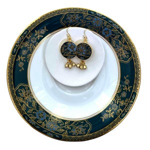 K&ouml;nnte beinhalten: Ein Paar goldene Ohrringe mit blauen und schwarzen Emaille-Blumenmustern und goldenen Perlen, die an der Unterseite h&auml;ngen. Die Ohrringe werden auf einem wei&szlig;en Teller mit einem gr&uuml;nen und goldenen Blumenmuster pr&auml;sentiert.
