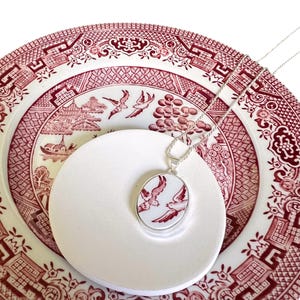 Puede incluir: Un collar con colgante de plata con un dije ovalado de porcelana que representa pájaros rojos. El colgante descansa sobre un plato blanco, sobre un fondo de un plato con estampado rojo y blanco. La cadena del collar es plateada.