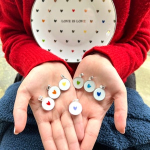 Könnte beinhalten: Ein Set aus sechs silbernen Anhängern mit bunten Herzmotiven. Jeder Anhänger hat einen weißen Hintergrund mit einem einzelnen Herz in einer anderen Farbe: Rot, Orange, Gelb, Grün, Blau und Lila.