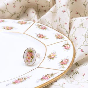 May include: An oval ring featuring a white ceramic face with a pink and yellow rose floral pattern. The ring has a metallic band and is positioned on a white porcelain plate with a matching floral border and gold trim detail.