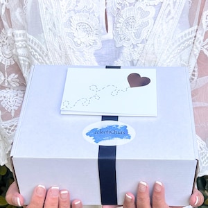 Puede incluir: Una caja de regalo blanca con una cinta negra y una tarjeta con un dise&ntilde;o de coraz&oacute;n. La caja tiene una pegatina con el texto "eclectiQuas". La caja es sostenida por una persona que lleva una blusa de encaje blanca.