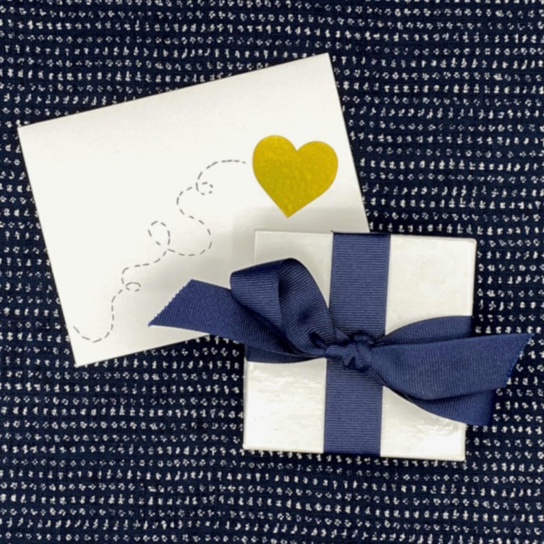 K&ouml;nnte beinhalten: Eine wei&szlig;e Geschenkbox mit einem dunkelblauen Band, das zu einer Schleife gebunden ist. Die Box steht auf einer blau-wei&szlig;en gemusterten Oberfl&auml;che. Hinter der Box liegt eine wei&szlig;e Karte mit einem goldenen Herzen und einer gestrichelten Herzzeichnung.