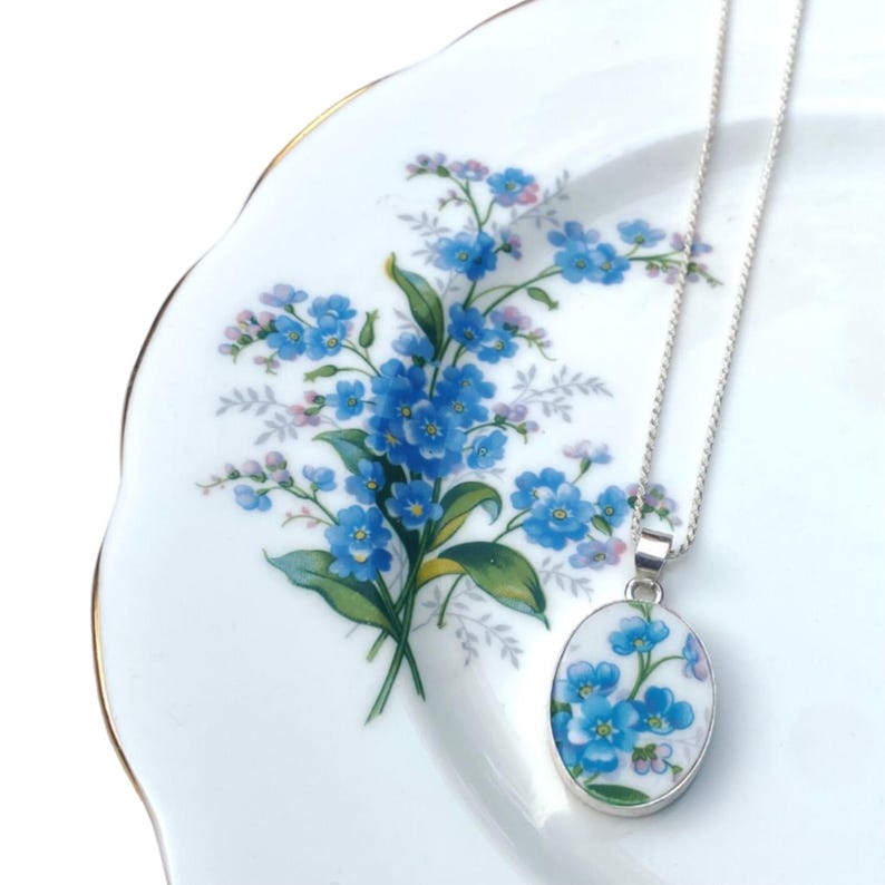 Pode incluir: Um colar de prata com um pingente oval com um design floral azul. O pingente pende de uma corrente delicada. O colar &eacute; exibido em um prato branco com um padr&atilde;o floral azul e acabamento dourado.
