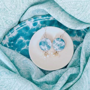 May include: Round earrings with a silver frame, displaying a blue and white pattern. Each earring has small, dangling beads. They are presented on a small, off-white plate, set against a backdrop of patterned blue and white fabric.