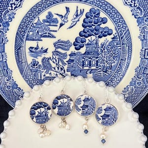 Puede incluir: Cuatro pares de pendientes redondos con un diseño de patrón Willow azul y blanco, cada uno con un gancho plateado. Algunos pendientes tienen colgantes de perlas o cristales azules. Los pendientes se exhiben en un plato blanco con un patrón Willow azul y blanco.