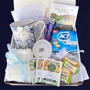 Puede incluir: Una caja de regalo con una taza floral, un cuenco peque&ntilde;o con una "M", un pa&ntilde;o blanco con una cinta azul, bolsitas de t&eacute;, pastillas y un libro titulado "Finding Inspiration in the Bird's Journey".