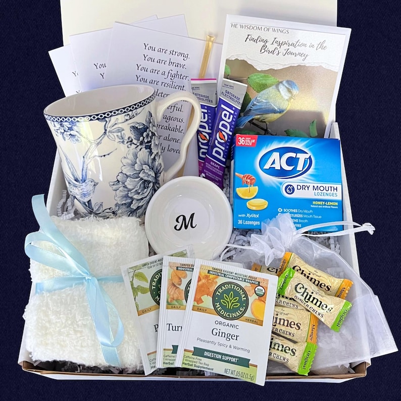 Puede incluir: Una caja de regalo con una taza floral, un cuenco peque&ntilde;o con una "M", un pa&ntilde;o blanco con una cinta azul, bolsitas de t&eacute;, pastillas y un libro titulado "Finding Inspiration in the Bird's Journey".