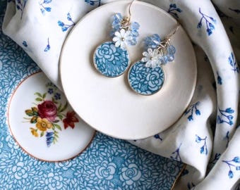 May include: A pair of round, blue and white floral earrings with gold-colored hooks. The earrings are adorned with small white flowers and blue beads. They rest on a white plate, with a floral patterned background.