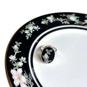 Può includere: Un anello d'argento con una pietra ovale nera che presenta un fiore bianco con foglie verdi. L'anello è su un piatto bianco e nero con un design floreale.