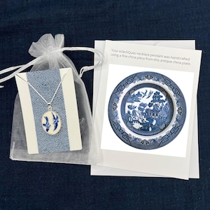 May include: A white organza bag with a blue and white porcelain pendant necklace inside. The pendant features a blue and white willow pattern. The bag is sitting on a white card with the text "Your eclectiQuas necklace pendant was handcrafted using a fine china piece from this antique china plate."