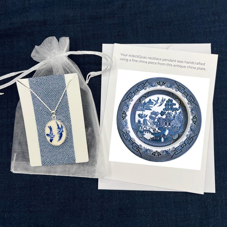 May include: A white organza bag with a blue and white porcelain pendant necklace inside. The pendant features a blue and white willow pattern. The bag is sitting on a white card with the text "Your eclectiQuas necklace pendant was handcrafted using a fine china piece from this antique china plate."